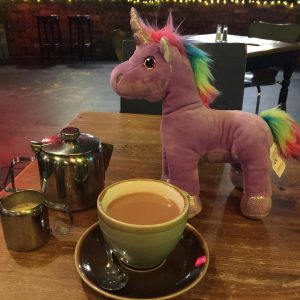 A picture of Steve the Unicorn - a purple plush unicorn with a rainbow mane and tail - sitting on a table with a cup of tea